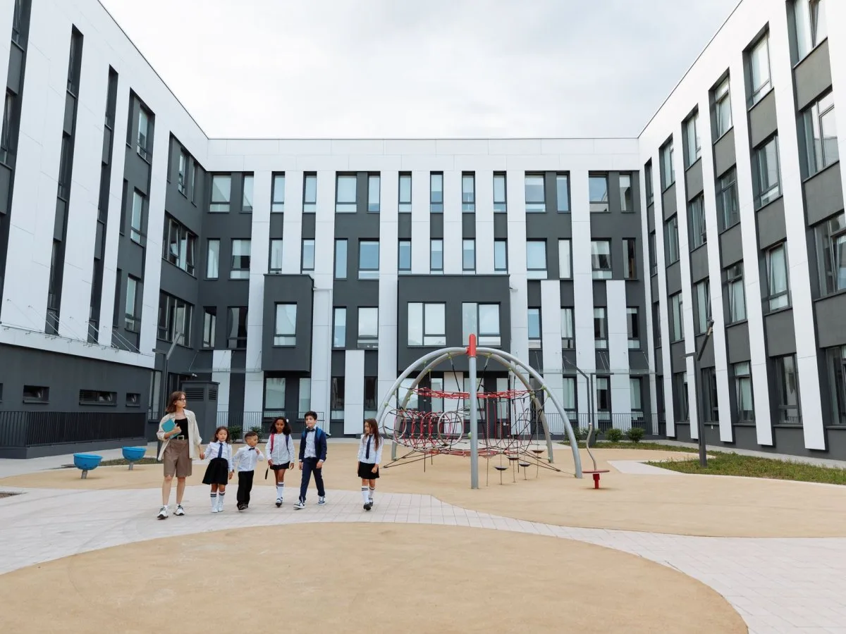 Eine Gruppe Kinder und eine erwachsene Person gehen an einem Spielgerät vorbei durch den Innenhof einer modernen Schule.
