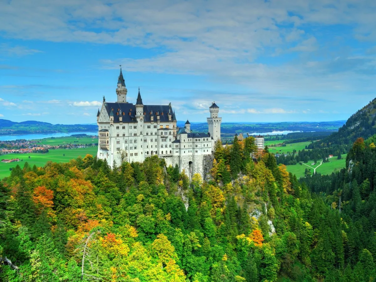 Le château de Neuschwanstein domine un versant boisé aux couleurs de l’automne, avec des champs et des montagnes au loin.