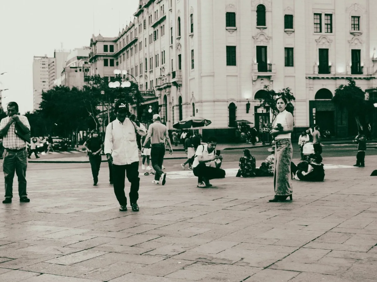 Persone di diverse età che camminano e si siedono in una piazza urbana pavimentata, circondata da edifici storici di più piani.