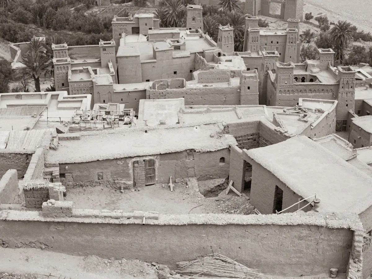 Black-and-white high-angle view of a mud-brick settlement with earthen walls, flat roofs, arched decorative towers and a grove of palm trees in the background.