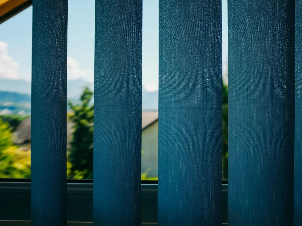 Vue à travers des stores verticaux bleus vers un paysage extérieur avec des arbres et un ciel dégagé.